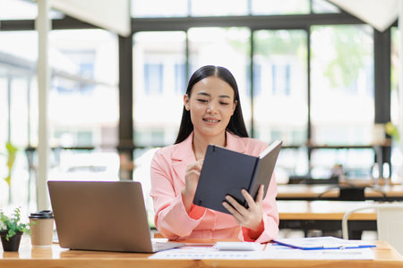 Asian businesswoman analyzing a report pointing to a graph with a pen, laptop and calculator. In workstation accountant telemarketing concept, ecommerce, free online marketing.の写真素材