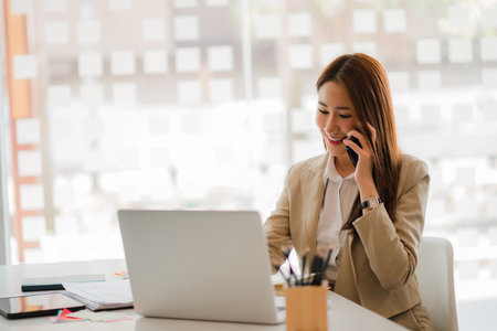 Young Asian business woman using smartphone to talk to customers, work with laptop and financial graphs to calculate, financial accounting, online work concept.の写真素材
