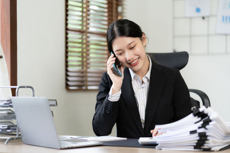 Young Asian female accountant with a smiling face as she uses her phone, computer and financial documents on her desk, planning concepts, analyzing market data.の写真素材
