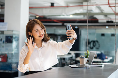 Attractive Asian woman talking on the phone and smiling while sitting at her office with laptop in the office.の写真素材