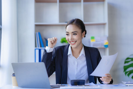 Beautiful Asian businesswoman sitting on laptop excited and happy with work accomplished. happy and satisfied with successful businessの写真素材