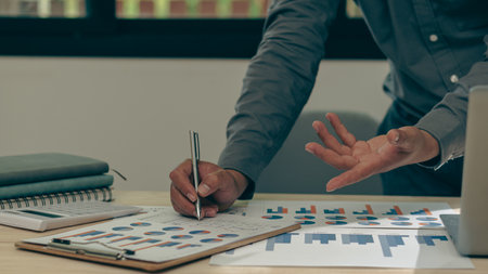 Close up of businessman hand holding pen and pointing at financial documents and investment plan for business and stocks and graph chart for investment analysis in his officeの写真素材