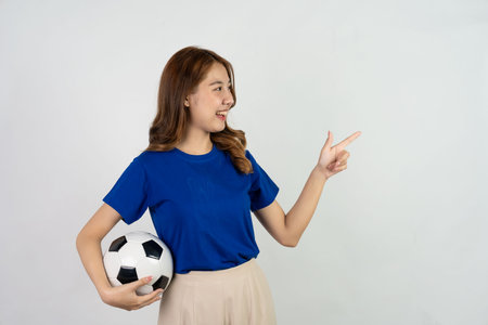 Happy Asian female soccer fan in blue shirt cheering for favorite team with soccer ball isolated on white background, pretty young pretty girl holding soccer ball, sports conceptの写真素材