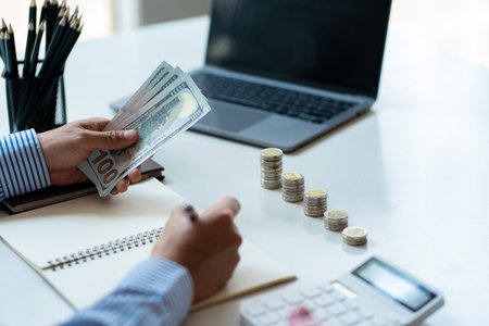Young man is using pen to record amount of money and coins for accounting plan on wooden table in home office, accounting and finance calculation conceptの写真素材