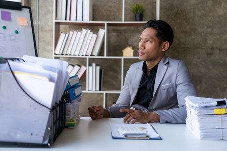 Young businessman sitting at desk with stack of papers facing window, business accounting conceptの写真素材