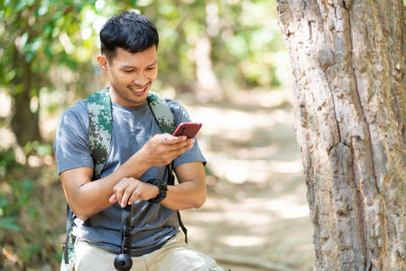 Handsome asian young male traveler with backpack, holding walking stick and hiking stick, standing in the forest with happiness, freedom in summer vacation, travel concept.の写真素材