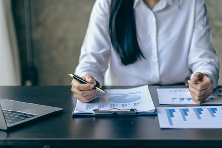 Close up of businesswoman holding pen pointing to paper, business chart for doing math on wooden table, tax, analytical accounting and calculating annual income and expenses, finanの写真素材