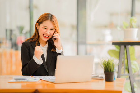 Smiling Asian woman talking on the phone with a customer Young positive female accountant using smartphone talking to team at her desk relaxing with mobile applicationの写真素材