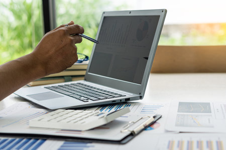 Businessman holding pen pointing to market graph and working on laptop computer working using calculator for mathematics on wooden table at office finance concept close up picturesの写真素材