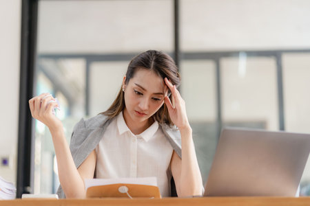 Thinking Asian businesswoman looking at financial statements and making marketing plan using computer on her desk in office holding report papers girl reading financial informationの写真素材