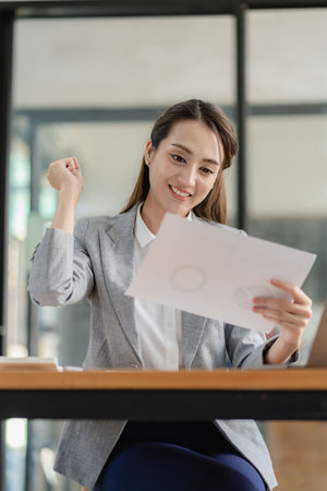 Happy young asian businesswoman celebrating success with arms raised in front of laptop with excitement at officeの写真素材