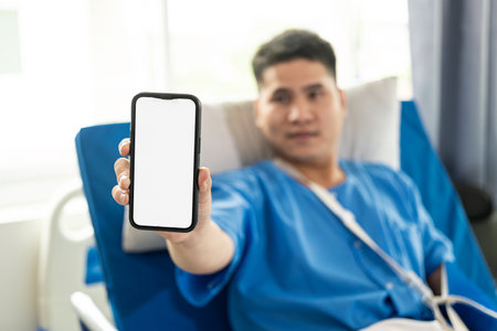 Asian male patient wearing sick clothes feeling happy holding blank white screen smartphone in hospital bed, health insurance conceptの写真素材