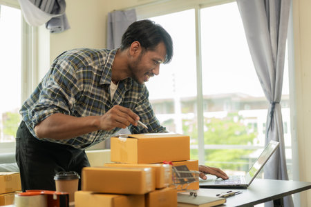 Receive orders via notebook to sell products online Ideas for online sales work at home, Startup, small business, SME. Handsome Asian guy preparing products for parcel boxes. Sellの写真素材
