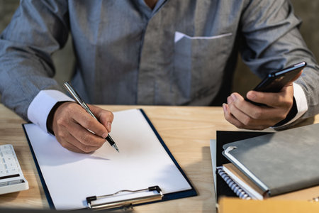 Man working at home holding pen checking business contract from document before signing to create new business SME. Close-up of businessman hand holding pen and using smartphone.の写真素材