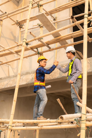Two Asian male engineers shaking hands and working together Hope stands on the architectural construction site on the structure industry. Wear a helmet with a checkered shirt.の写真素材