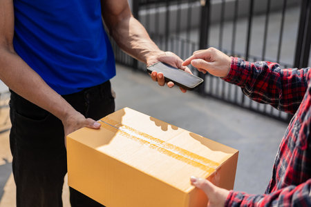 Smiling african american courier holding cardboard box delivering parcel to business customer signing electronic signature POD device on smartphoneの写真素材