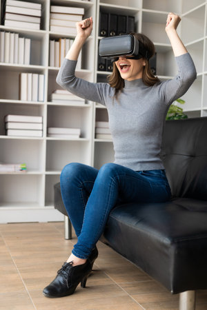 Happy young woman surprised in VR headset, feeling air. Smiling caucasian woman wearing vr glasses, virtual reality technology device, watching 3d simulation video while sitting onの写真素材