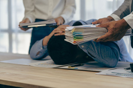 A male employee who is stressed and tired from working with a pile of documents and his co-worker brings a large amount of unfinished documents to him.の写真素材