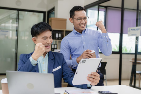 Two Asian businessmen raising their hands to celebrate success, rejoicing in increasing profits in business are getting good news online.の写真素材