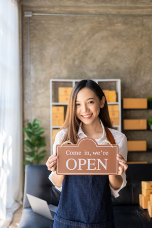 Happy young business owner standing holding open sign Happy Asian woman wearing an apron hanging a...
