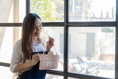 Happy young business owner standing holding open sign Happy Asian woman wearing an apron hanging a...