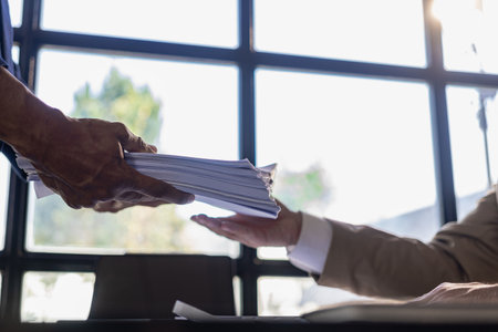 Businessman's hand holding a pile of papers to search for information about his home office on his desk. Business report. A pile of unfinished documents lies on the table.の写真素材