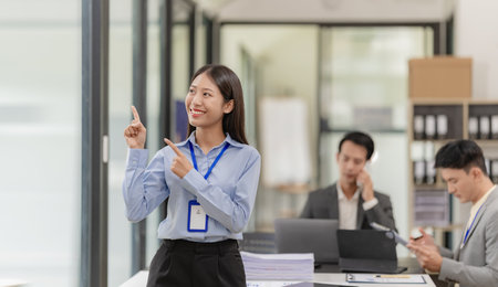 A female Asian accountant stands in front of a desk and two businessmen meet to discuss brainstorming, calculations, and joint analysis of the company's growth goals.の写真素材