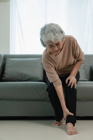An elderly Asian woman grimaced in pain in her right ankle. Sitting down on the sofa, having an injury from walking too much.の写真素材