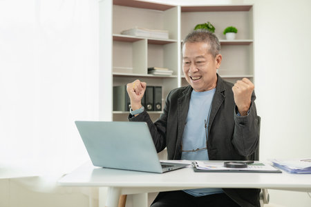 Senior Asian businessman in a suit is excited Happy mature Asian businessman clenching his fist showing victory gesture while sitting on desk in front of laptop at officeの写真素材