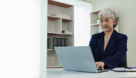 Senior Asian businesswoman working with graph documents to analyze finances and work sitting at table in office, typing on keyboard, studying financial documents, analyzing profitの写真素材
