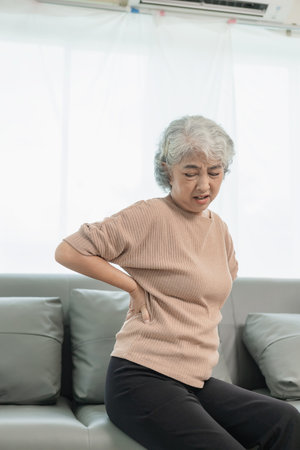 An elderly woman has back pain after waking up while sitting on the sofa at home. An elderly Asian...