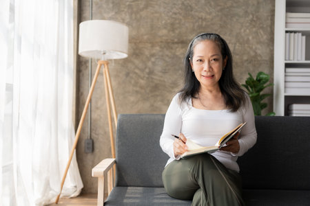 Mature Asian woman sitting on sofa and writing in notebook at homeの写真素材