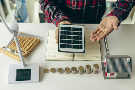 Professional male green energy engineer sitting at a work desk with solar panels and wind turbines, young man recommends ways to save electricity and money.の写真素材