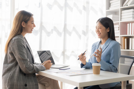 Two young Asian businesswomen work on laptop computers in the office with reports. Colleagues discuss financial data in document analysis of new marketing projects.の写真素材