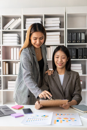 Two young Asian businesswomen work on laptop computers in the office with reports. Colleagues discuss financial data in document analysis of new marketing projects.の写真素材