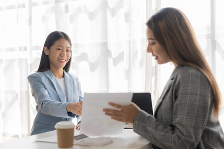 Two young Asian businesswomen work on laptop computers in the office with reports. Colleagues discuss financial data in document analysis of new marketing projects.の写真素材