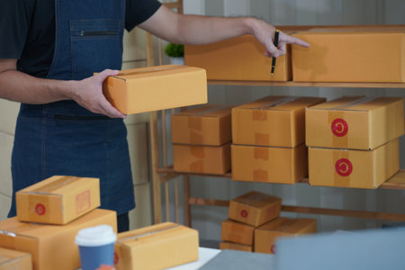 SME business Young man working with laptop with cardboard boxes stacked on table ready for delivery,...