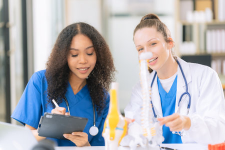 Two beautiful female doctors sitting at a table in a science lab with test flasks. Colorful glass bottles filled with various liquids on table with microscopeの写真素材