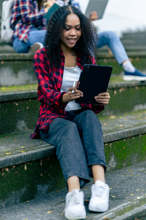 Diverse young university students working outdoors using laptops and tablets together discussing technology learning concepts.の写真素材