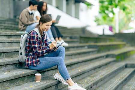 Diverse young university students working outdoors using laptops and tablets together discussing technology learning concepts.の写真素材