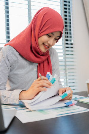Muslim female accountant wearing hijab holding pen and working with calculator to calculate business data, accounting documents and laptop computer in office. Vertical close-up phoの写真素材