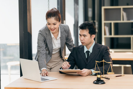 Lawyers discuss contract documents at a wooden table. Symbols of justice are hammer and scales,...
