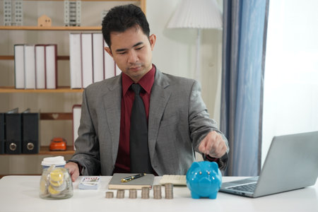 Man in suit using modern laptop and stack of coins with blue piggy bank on his desk. Business,...