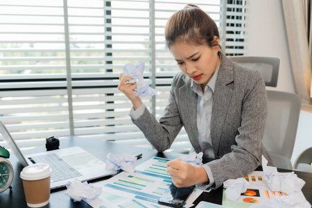 Asian businesswoman stressed holding crumpled paper while working with laptop and documents in...