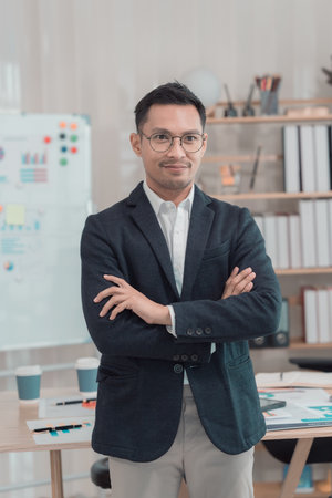 Image of young Asian businessman standing in office with arms crossed, confident standing in front...