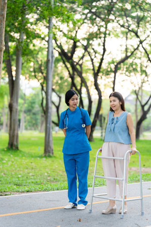 Asian nurse assists elderly woman who walks with a cane. Assists and cares for elderly female patient who is doing physical therapy in a park.の写真素材