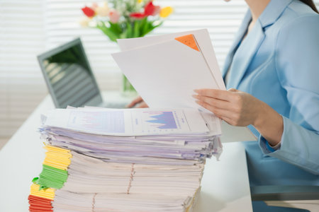 Close-up of businesswoman holding a pile of documents at her desk. Documents are neatly arranged on her desk, possibly preparing a report or document.の写真素材