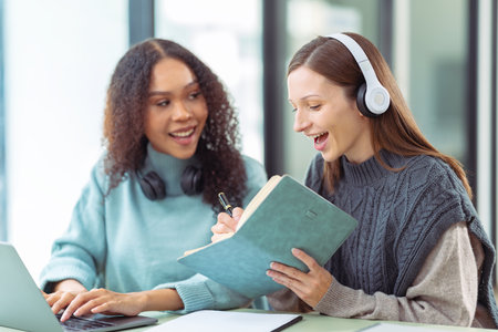 Two young female students wearing headphones studying using laptops for video chat. Millennial...