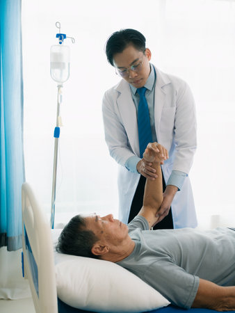 A skilled male physical therapist examines an elderly male patient lying on a bed, checking his...