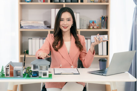 Real estate agent sitting in office workplace, concept of buying and loan in home title working, laptop computer while other hand holding model house.の写真素材